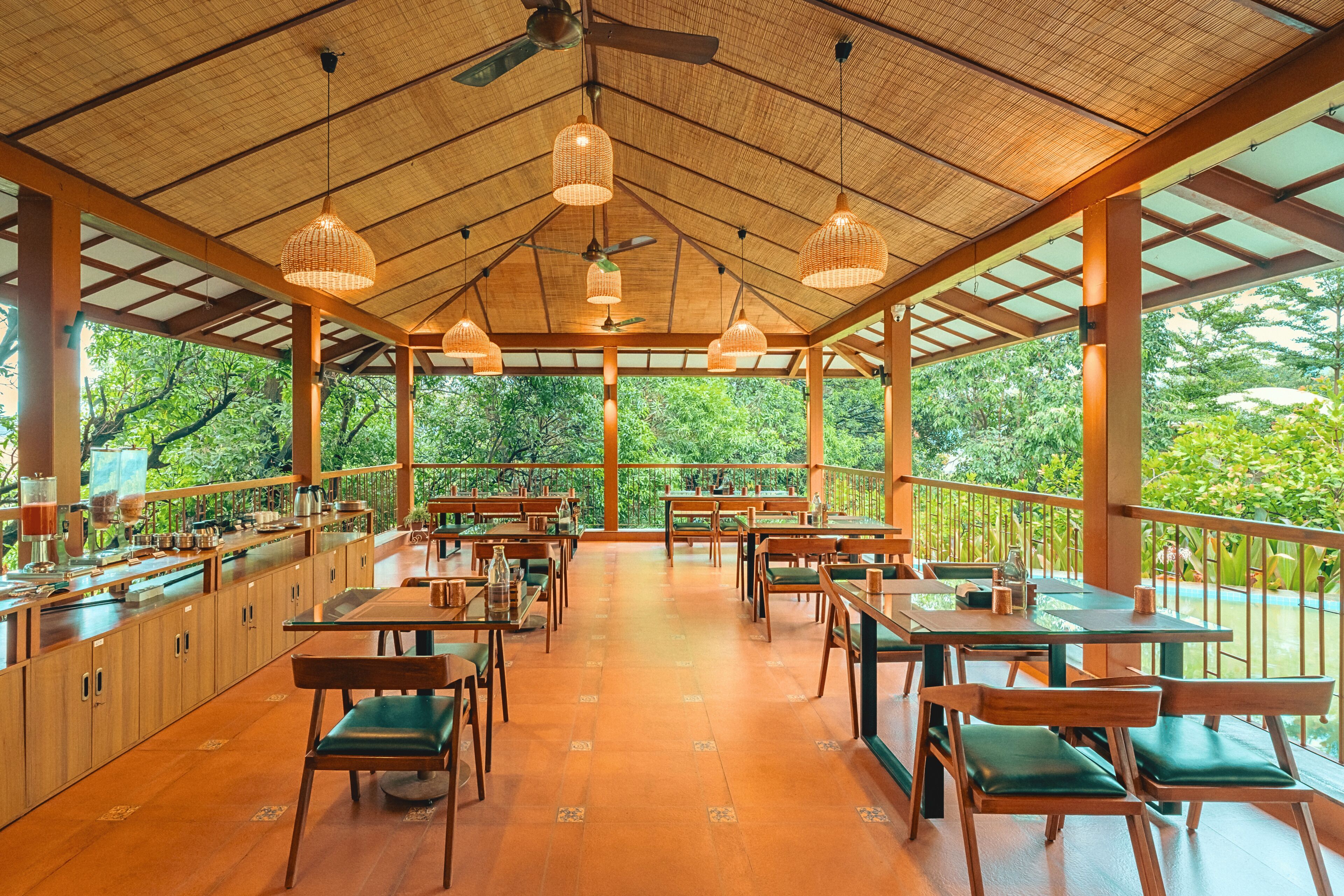 An open-air restaurant with wooden tables and chairs, a bar area, and a view of lush greenery.