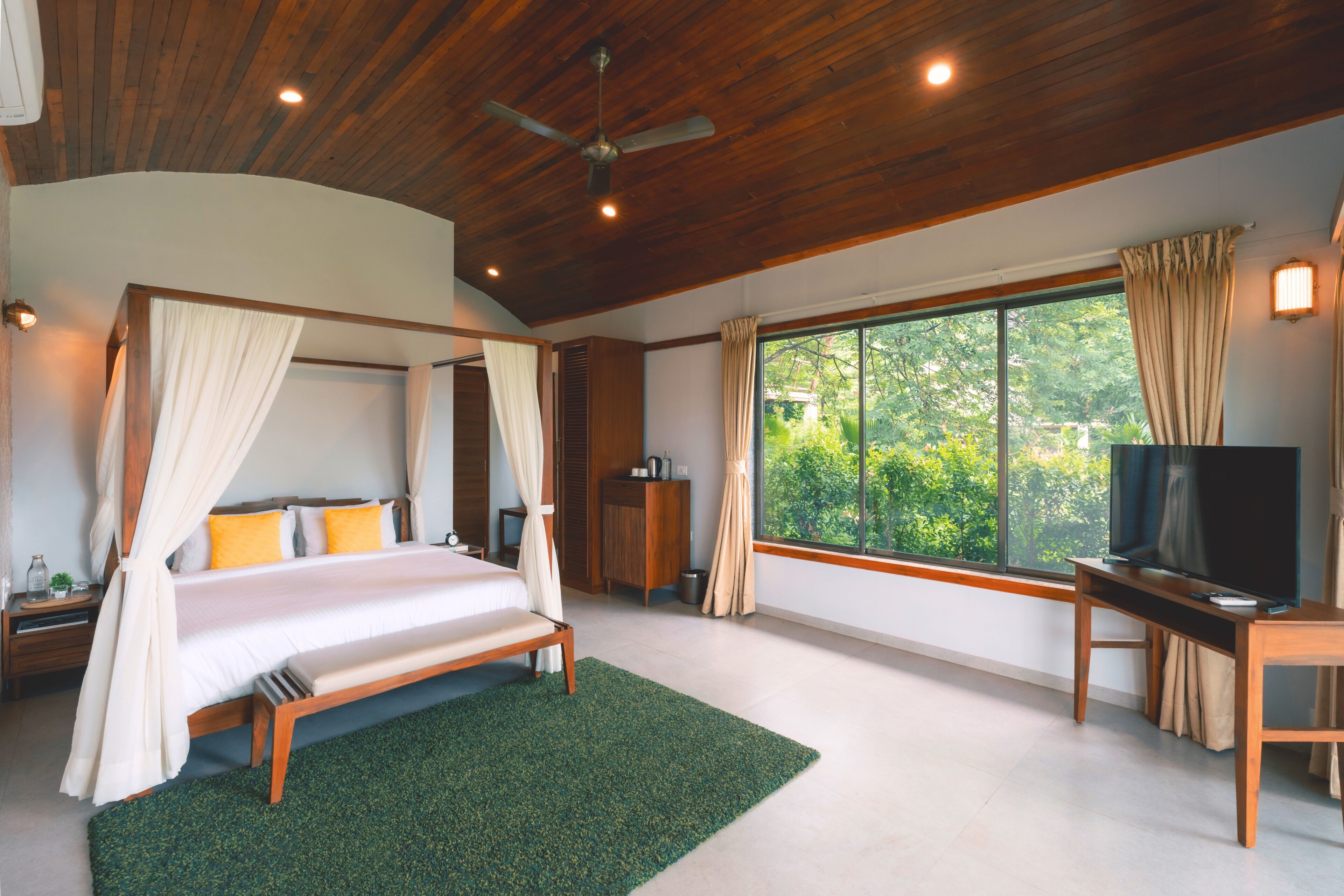 A bedroom with a four-poster bed, a television, a desk, and a large window with curtains.