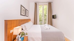 A hotel room with a wooden headboard, a bed with white linens, a bedside table with a lamp, a TV mounted on the wall, and a window with curtains.