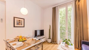 A modern living room with a dining area, a flat-screen TV, a large window with curtains, and a potted plant.