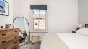Une chambre à coucher avec un lit, une commode en bois, un miroir, une fenêtre donnant sur un immeuble en briques et un tableau encadré accroché au mur.