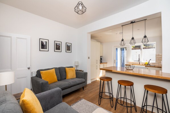 A modern living room with a grey sofa, yellow cushions, and a wooden coffee table. There are three bar stools and a kitchen area with a wooden countertop and hanging lights.
