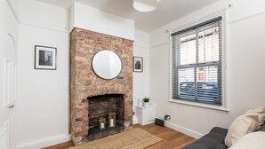 Un salon avec une cheminée en briques, un grand miroir rond au-dessus, un canapé gris et une fenêtre avec des stores.
