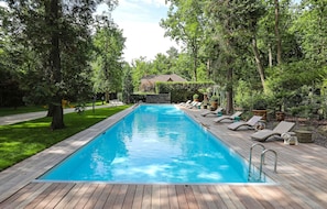 Una piscina rettangolare con un solarium in legno, circondata da vegetazione lussureggiante e alberi.