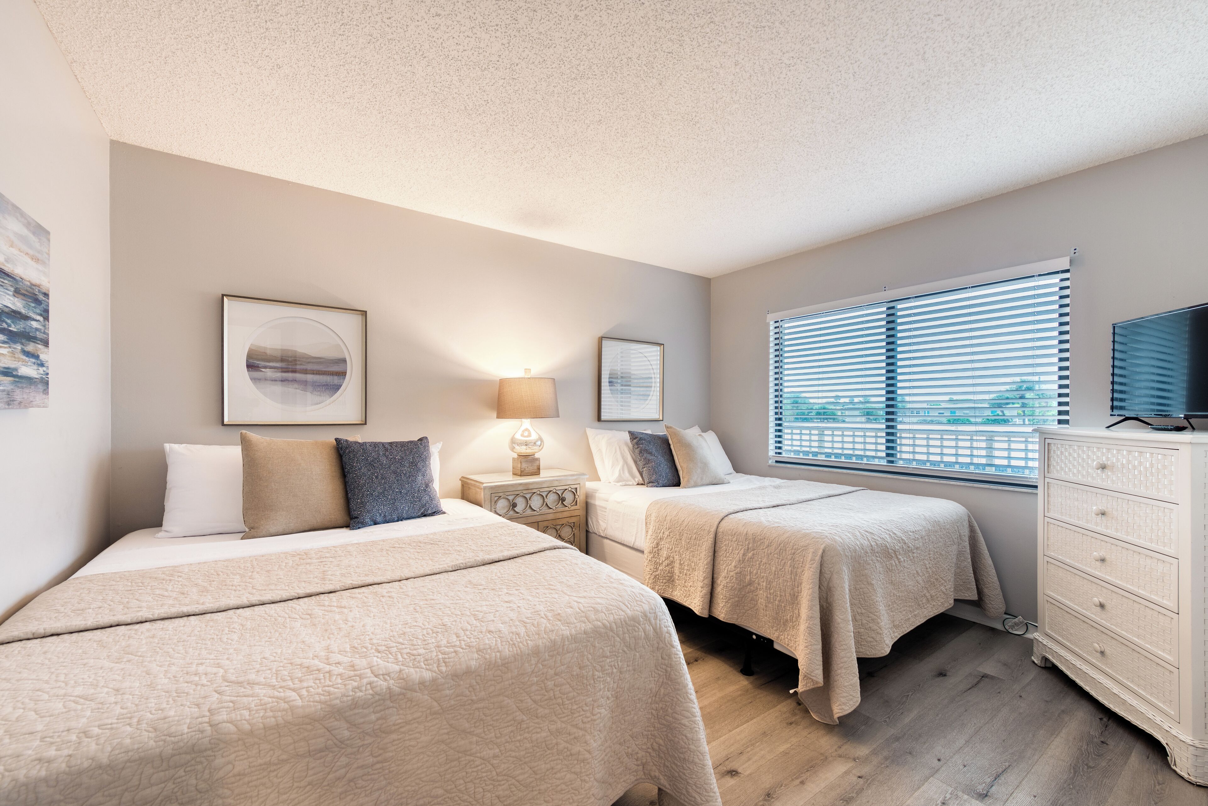 A bedroom with two beds, a dresser, a lamp, and a window with blinds.