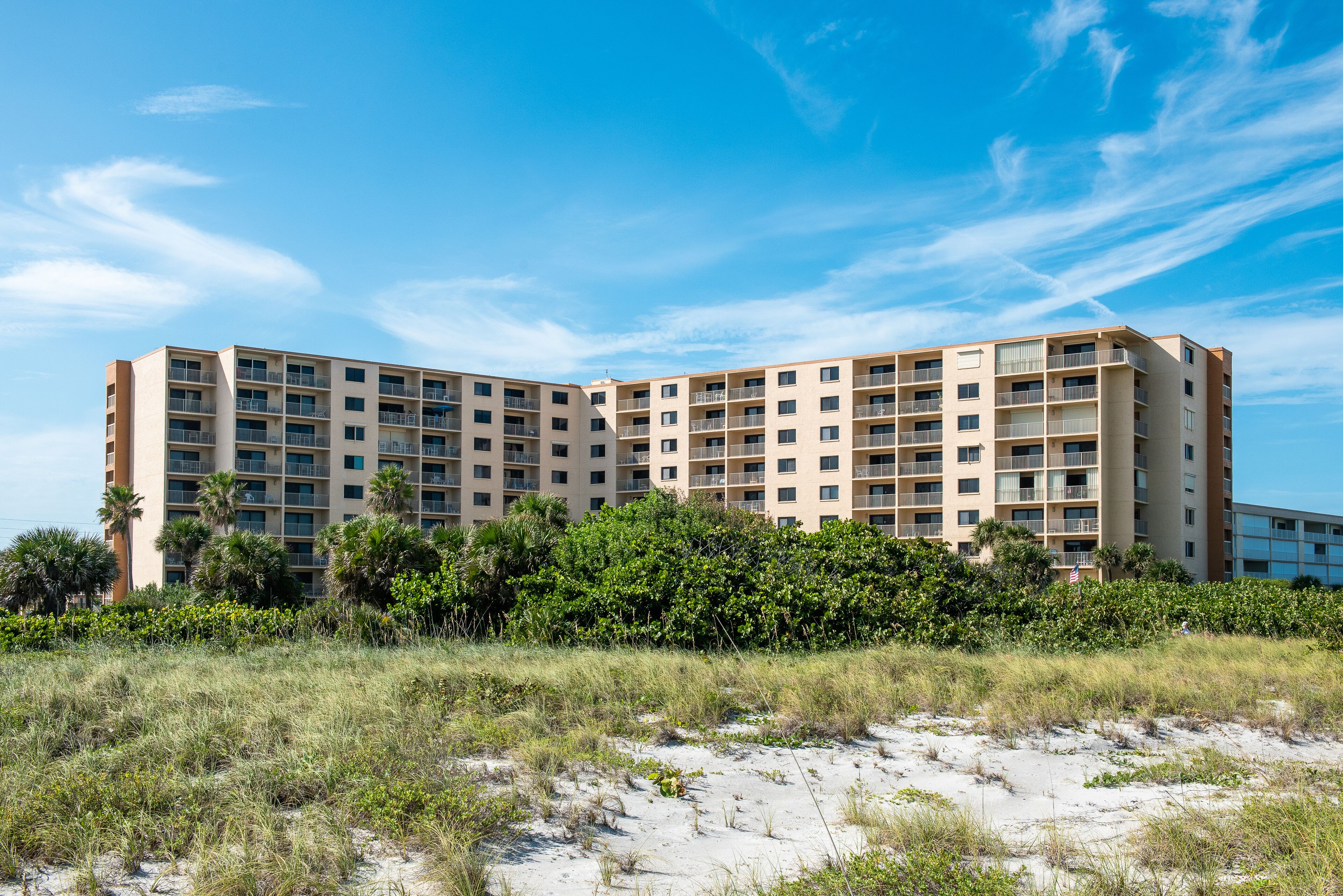 A multi-story hotel with balconies, surrounded by greenery and sand.