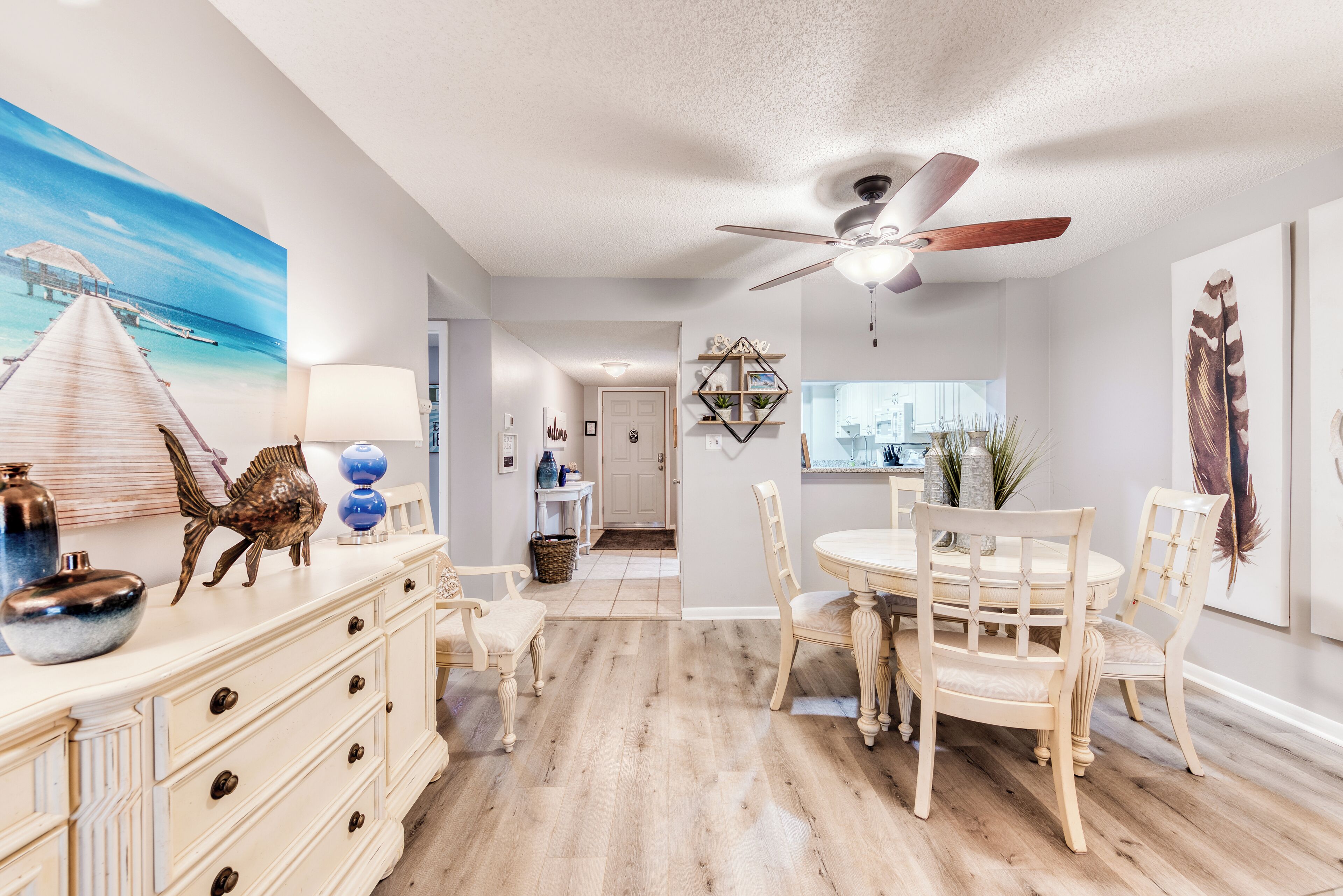 A dining area with a wooden table and chairs, a ceiling fan, and a painting of a beach scene.