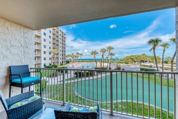 A balcony with wicker furniture, a view of a pool, and a building in the background.