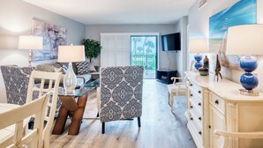 Un salon comprenant une table à manger, des chaises, un canapé et un téléviseur. Une fenêtre offre une vue sur les palmiers.