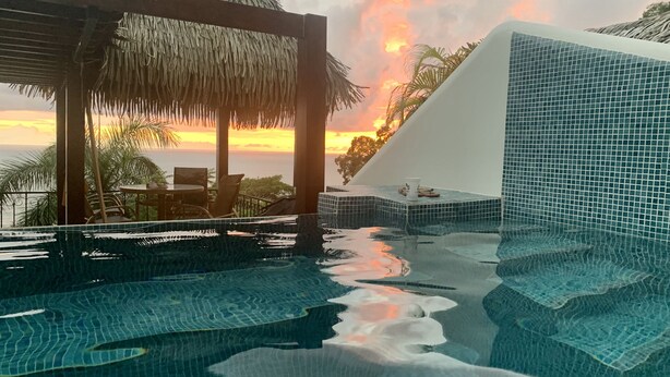A infinity pool with a view of the sunset, a thatched gazebo, and a tiled wall.