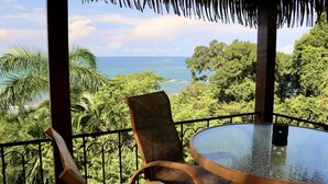 A glass table with chairs on a balcony overlooking a tropical landscape with a view of the ocean.