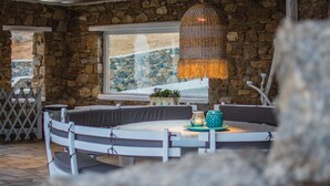 A round dining table with white chairs, a hanging woven light fixture, and a stone wall backdrop. -  6Bedroom Villa with private pool, located in Kalafatis, Mykonos (Καλαφάτη, Μύκονος)