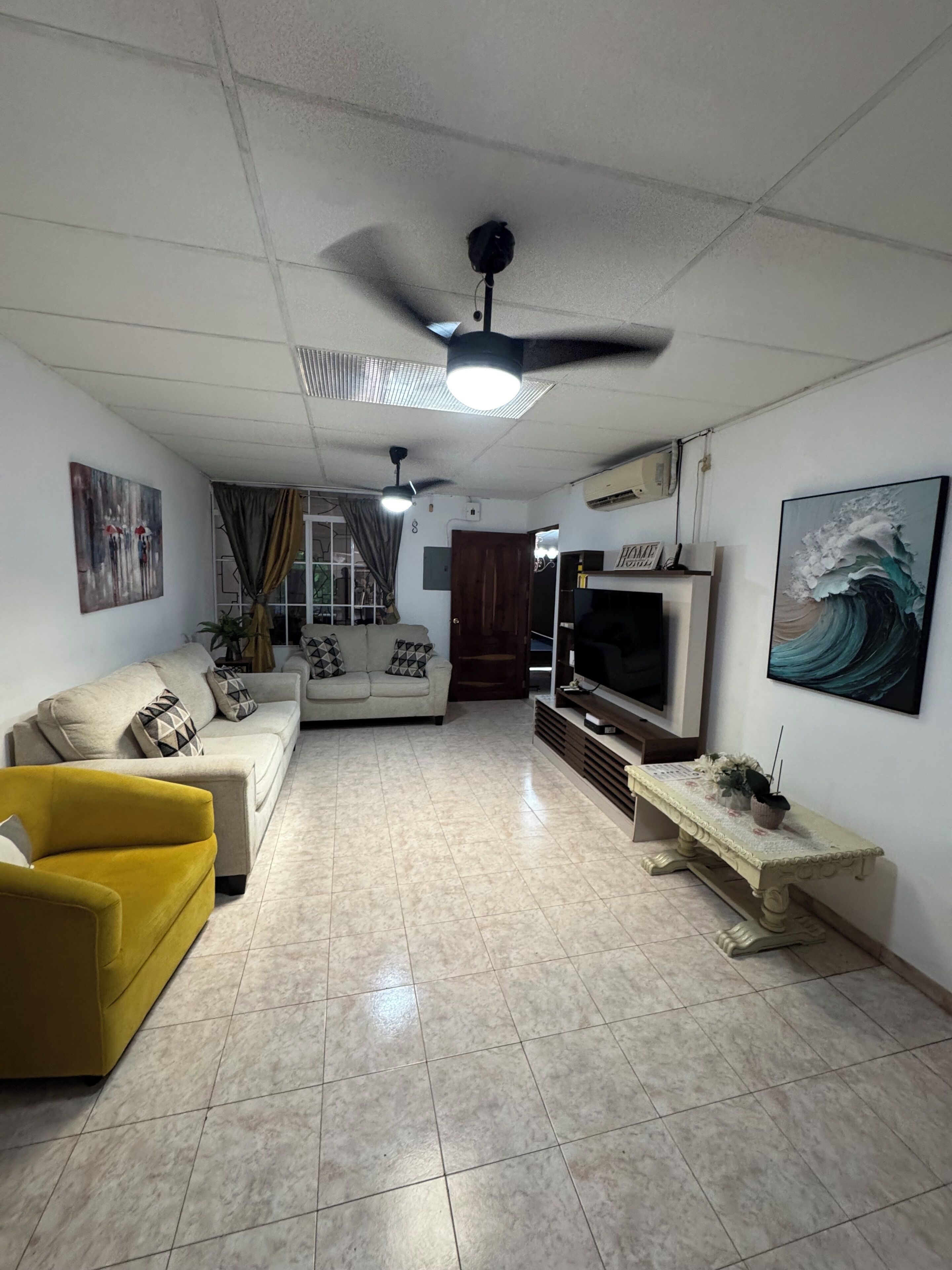 Un salón con sofá, un sillón amarillo, televisor y ventilador de techo.