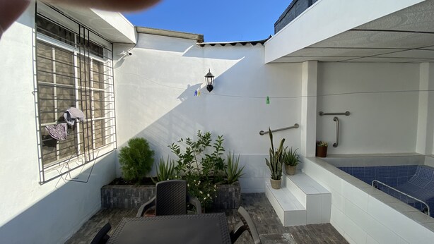 Une terrasse avec une table et des chaises, un petit jardin agrémenté de plantes en pot, une fenêtre avec un grillage métallique et un mur blanc avec un sol carrelé.