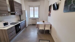 A modern kitchen with a microwave, stove, and sink. There is a dining table with chairs and a small potted plant. A radiator is under a window with white curtains. - Residence Bonomelli apt. no. 18 two-room apartment (Bergamo)