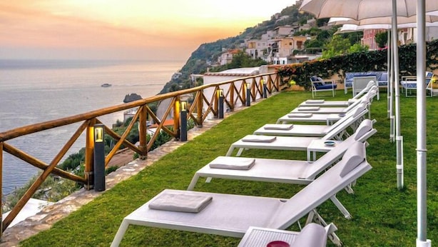 Une terrasse en bord de mer, avec des chaises longues blanches et des parasols, offrant une vue sur l’océan et une ville côtière.