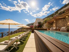 A poolside area with lounge chairs and umbrellas, a stone wall, and a view of the sea.
