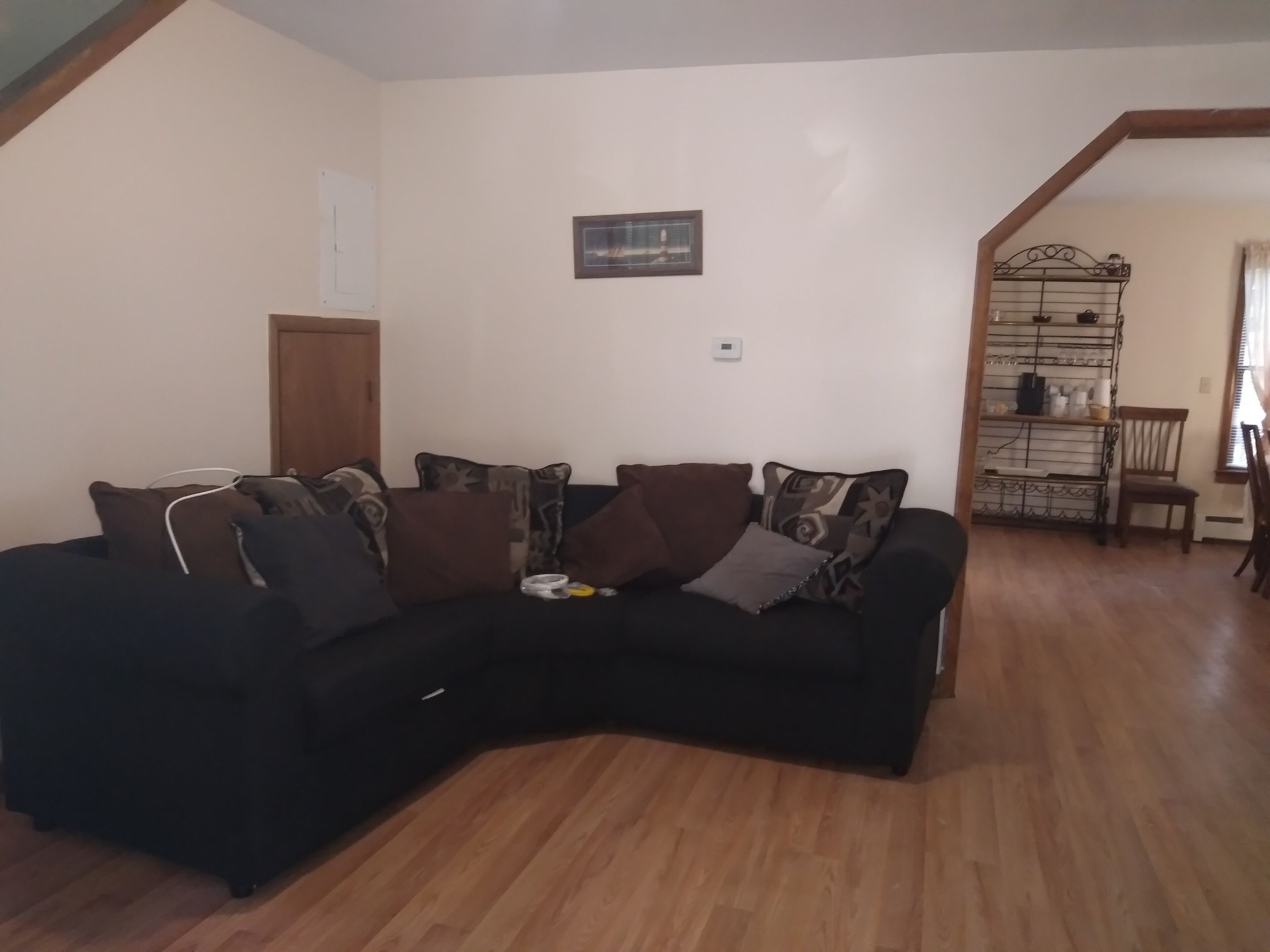A living room with a black sectional sofa, a wooden floor, a white wall, a door, and a metal shelf unit.