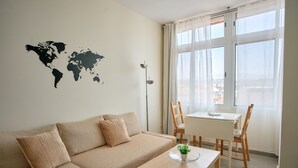 A living room with a beige sofa, a small table with a plant, a world map on the wall, and a window with curtains.