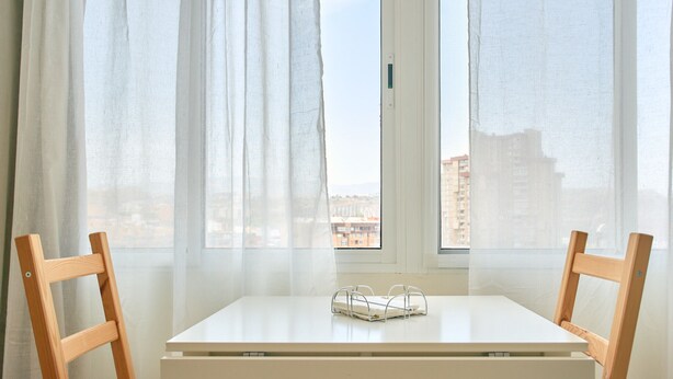 A room with a white table and two wooden chairs, a window with sheer curtains, and a view of buildings outside.