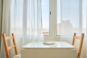 Una habitación con una mesa blanca y dos sillas de madera, una ventana con cortinas traslúcidas y vistas a edificios.