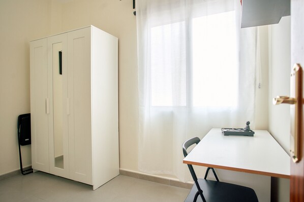 Una habitación con un armario blanco, una mesita con una silla y una ventana con cortinas de encaje.