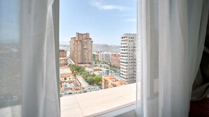 A view from a window with sheer curtains, overlooking a cityscape with multiple high-rise buildings and a clear sky.