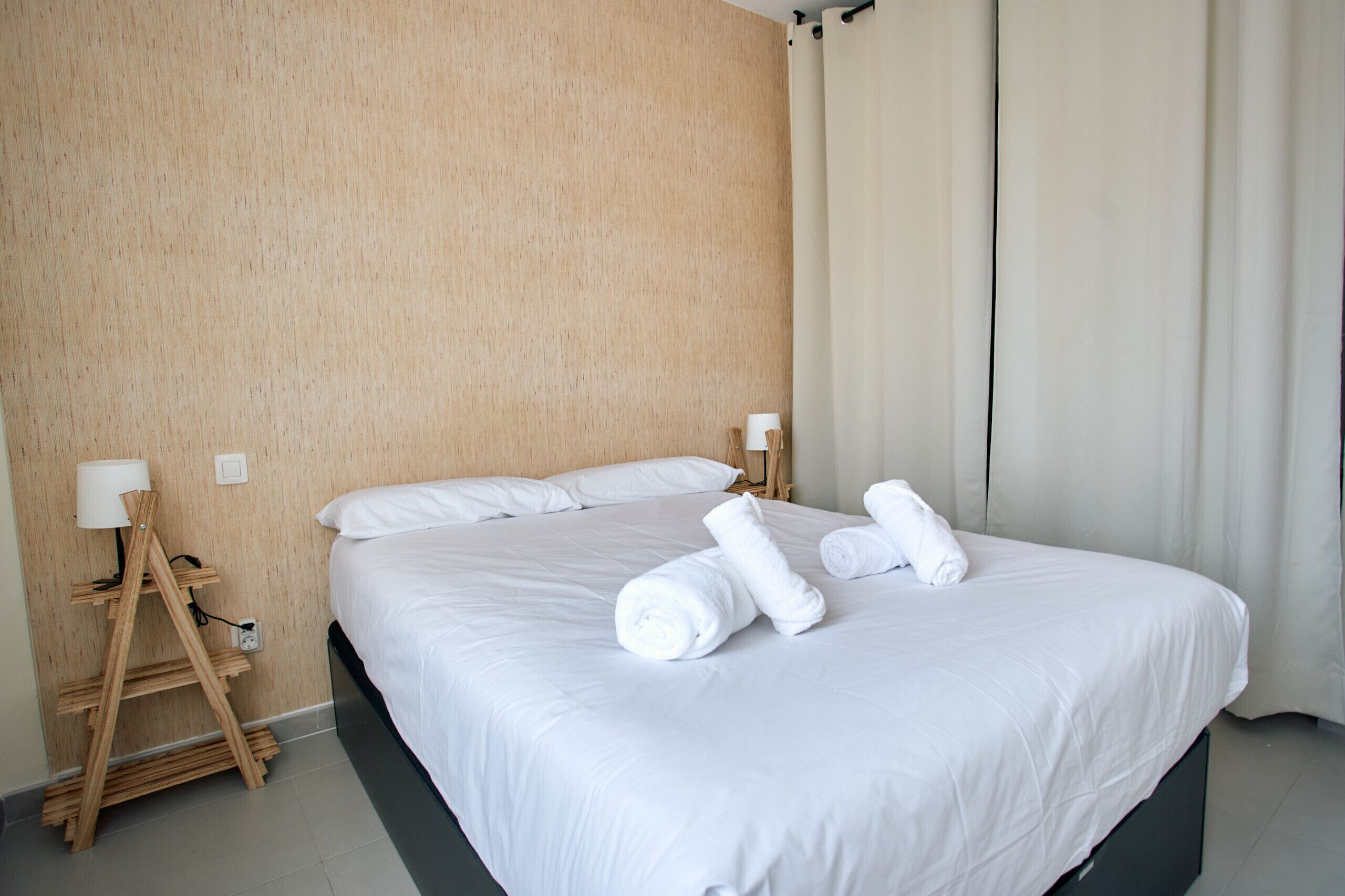 Una cama bien hecha con sábanas blancas y toallas enrolladas, una mesita de noche con lámpara y una estantería de madera apoyada en una pared con textura.