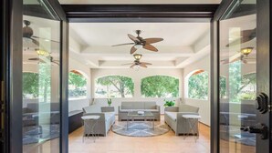 An open-plan living area with a sofa set, coffee table, and chairs, a ceiling fan, and large windows with a view of greenery. - Spacious Luxury Retreat Sleeps 20 with Jacuzzi (Fallbrook)