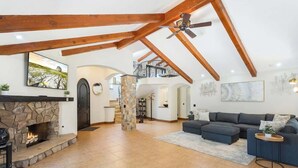 A spacious living room with a stone fireplace, a dark-colored sofa set, and a high ceiling with exposed wooden beams. - Spacious Luxury Retreat Sleeps 20 with Jacuzzi (Fallbrook)