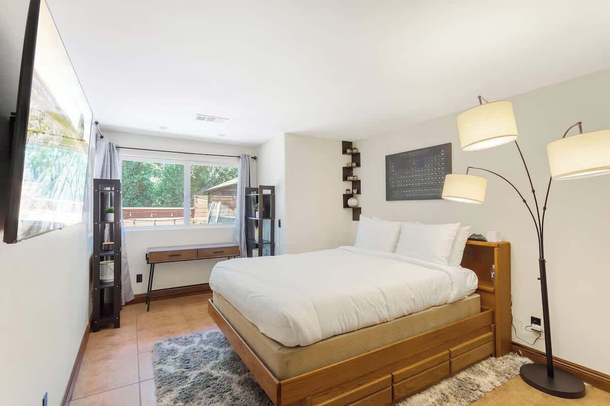 A bedroom with a wooden bed, a bench, a floor lamp, and a window with a view of greenery.
