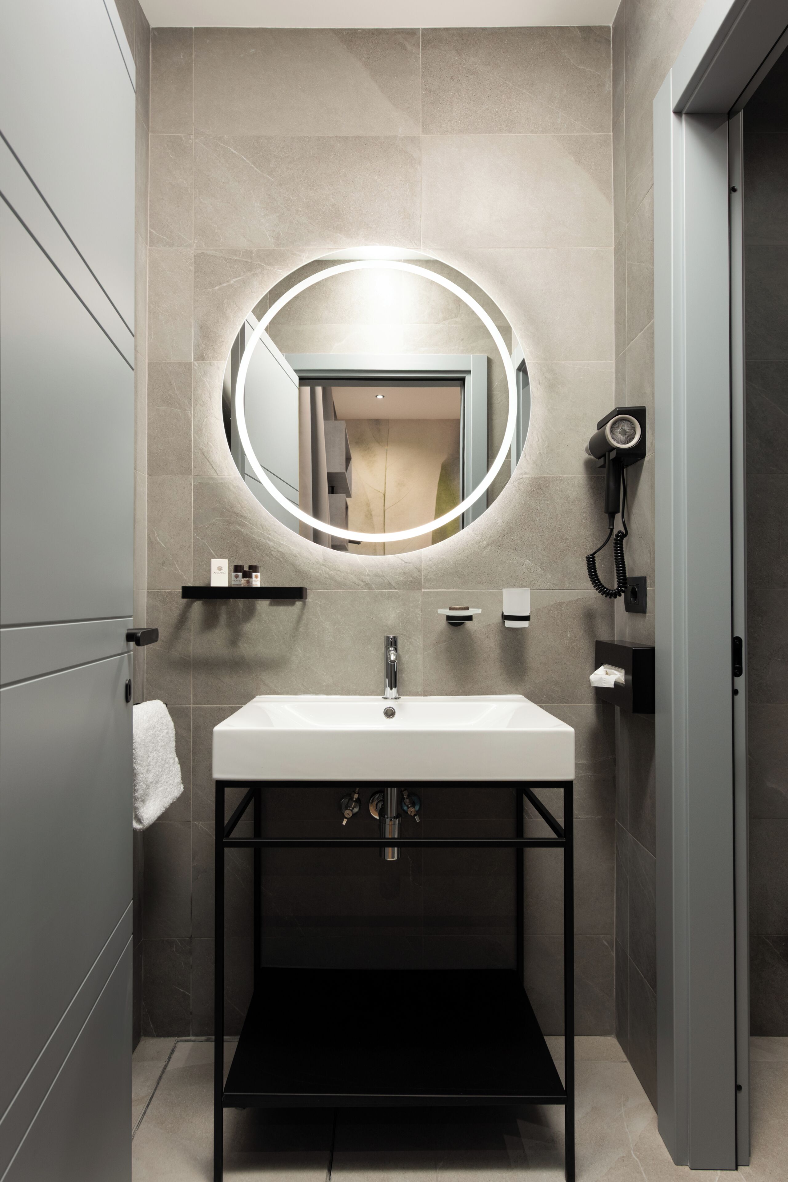 A modern bathroom with a round mirror, a white sink, and a hairdryer mounted on the wall.