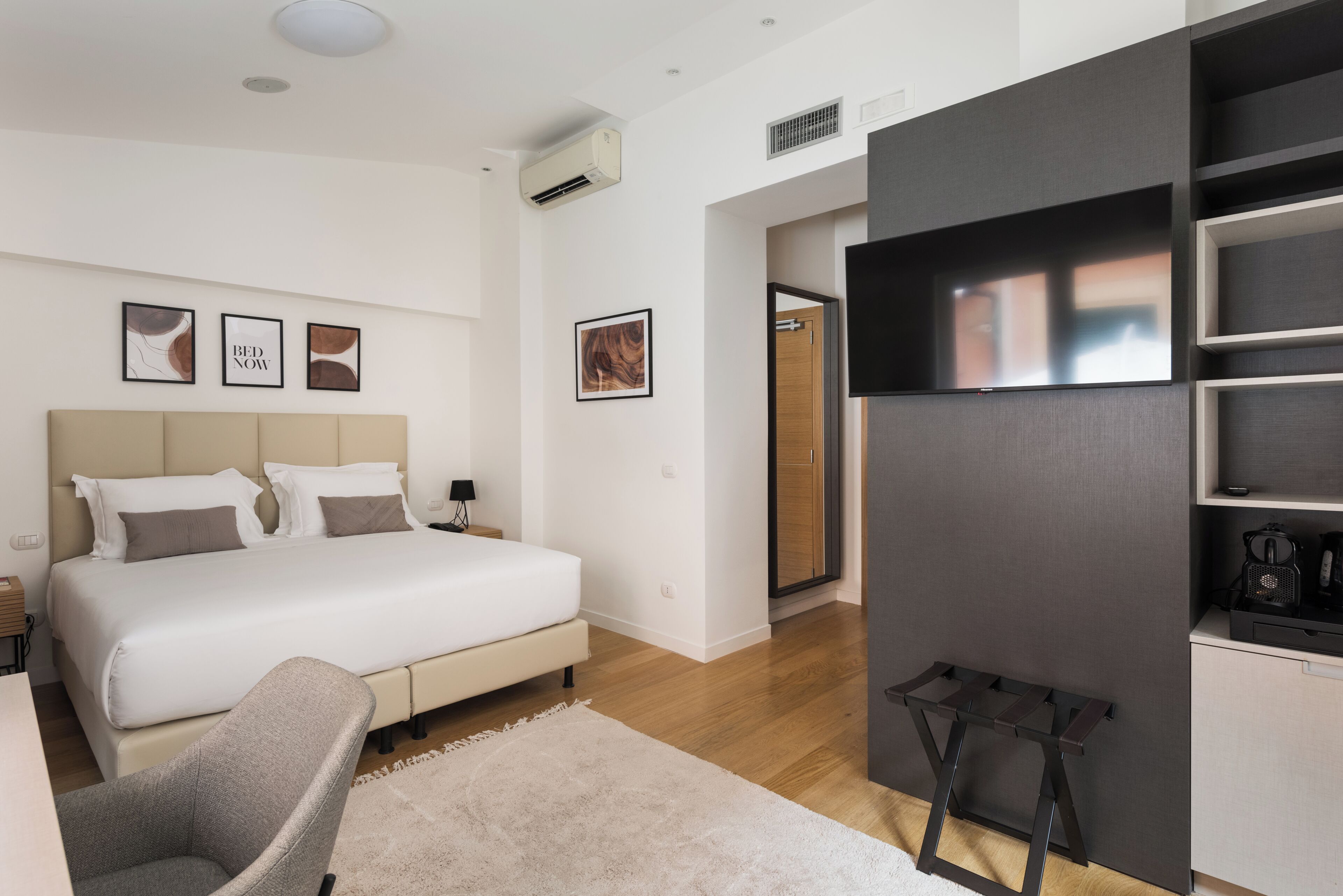 A modern bedroom with a large bed, a desk, and a chair. There is a TV mounted on the wall, a wardrobe, and a door leading to another room.