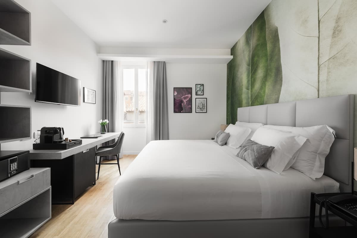 A modern hotel room with a large bed, a desk with a flat-screen TV, a coffee machine, and a view of the city through the window.