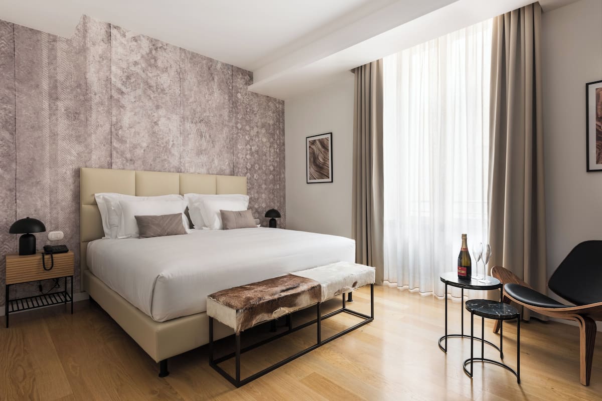 Une chambre d’hôtel moderne avec un grand lit, une banquette, une petite table avec une bouteille et une chaise.