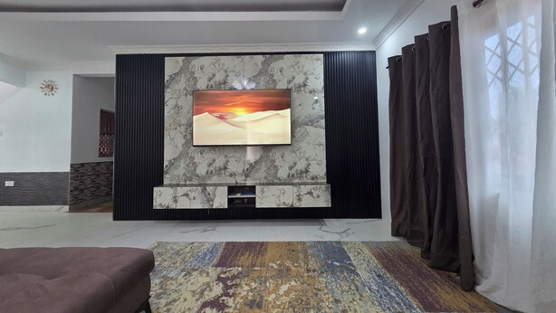 A modern living room with a flat-screen TV mounted on a marble wall, a patterned rug, a sofa, and a window with curtains.