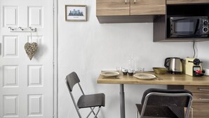 Eine kompakte Küche mit einer Mikrowelle, einem Toaster und einer Kaffeemaschine auf der Arbeitsfläche. Zwei Stühle und ein kleiner Tisch, an der Wand hängt ein Bilderrahmen.