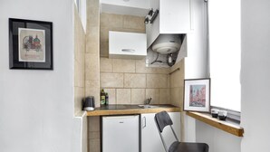 Eine kleine Küche mit Spüle, kleinem Kühlschrank und einem Stuhl. An der Wand befinden sich Bilder in Rahmen.