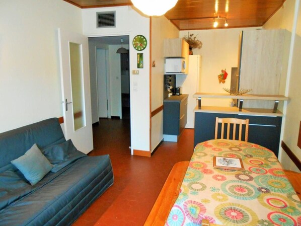 A compact room with a sofa, a dining table with a colorful tablecloth, a kitchenette, and a door leading to another room.