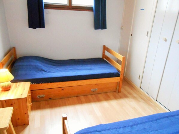 A small, single-bed room with a wooden bed frame, a wooden nightstand with a lamp, a small desk, and a wardrobe.