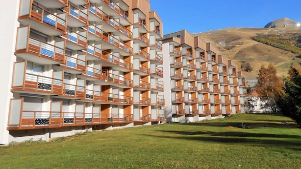 A multi-story hotel with balconies, a grassy courtyard, and a mountainous backdrop. - 50m from resort center, Ground floor, view ski slopes, balcony, parking, tv, ski locker, 30m² (Les Deux Alpes)