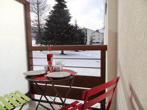A balcony with a table set for two, a red chair, and a view of a snowy landscape with trees and buildings in the background.