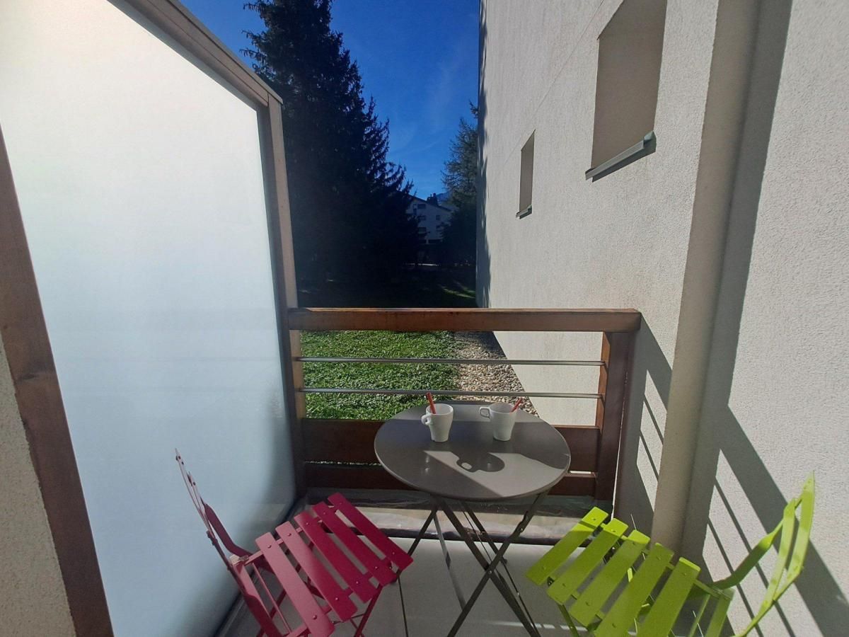 Ein kleiner Balkon mit Tisch und zwei Stühlen, eine weiße Tür sowie Blick auf eine grüne Wiese und Bäume.