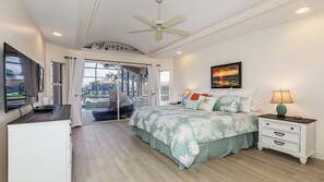 A bedroom with a bed, a nightstand, a TV, and a window with a view of a swimming pool and buildings.