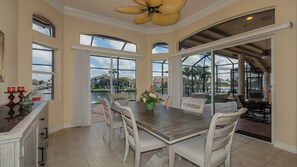 A spacious dining area with a large table and chairs, large windows offering views of a water body and buildings, and a ceiling fan.