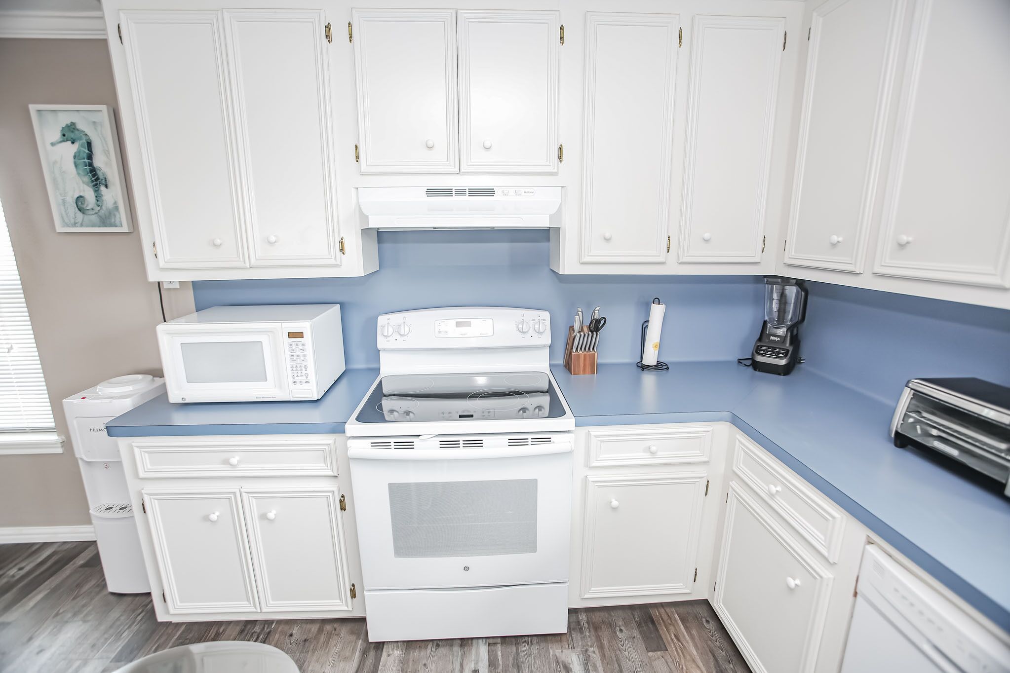 Una cocina con armarios blancos, microondas, horno, tostadora y licuadora sobre una encimera azul.