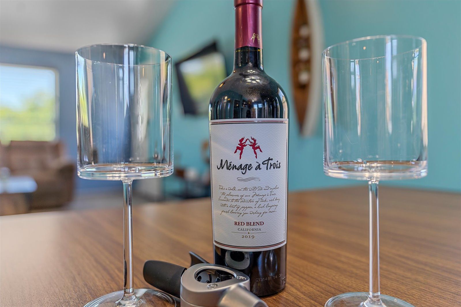 A bottle of wine with a label, two empty wine glasses, and a corkscrew on a wooden table.
