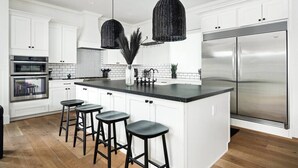 Una cocina moderna con armarios blancos, una isla grande con taburetes de bar, electrodomésticos de acero inoxidable y luminarias colgantes de fibra negra.