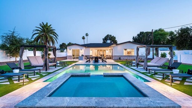 Una piscina rectangular con sillas de descanso y sombrillas, rodeada por un césped cuidado y una casa al fondo.