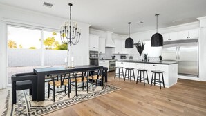 Una cocina moderna con armarios blancos, una isla grande con taburetes de bar, una lámpara de araña y un refrigerador de acero inoxidable.